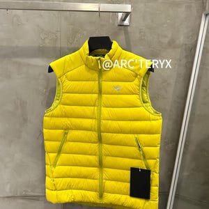 May include: A bright yellow puffer vest with a high collar and a full-length zipper. The vest has two side pockets and a small logo on the chest. The text "@ARC'TERYX" is visible on the top. The vest is hanging on a black hanger.