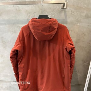 Op de afbeelding: Roestkleurige jas met capuchon die aan een metalen rek hangt. De jas heeft een capuchon en een ritssluiting. De merknaam "ARC'TERYX" is linksonder zichtbaar. De jas is gemaakt van een lichtgewicht materiaal.