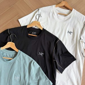 May include: Three short-sleeved t-shirts in black, white, and pale blue, each on a wooden hanger. They feature a small logo on the chest and contrasting stitching. The shirts are displayed on a light brown wooden surface. The shirts are made of a soft material.