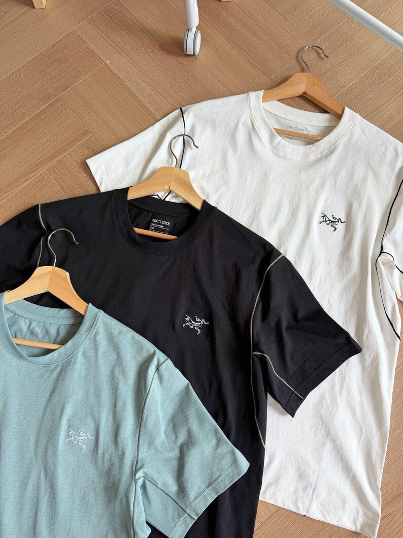 May include: Three short-sleeved t-shirts in black, white, and pale blue, each on a wooden hanger. They feature a small logo on the chest and contrasting stitching. The shirts are displayed on a light brown wooden surface. The shirts are made of a soft material.