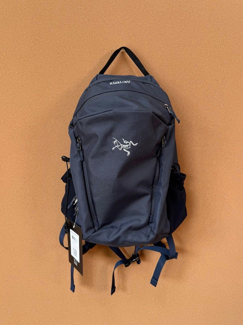 May include: A navy blue backpack with a black handle and straps. The backpack has a white logo and the word "ARCTERYX" printed on the top. It features multiple zippered compartments and adjustable straps.