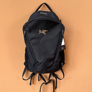 May include: Black backpack with a top handle and adjustable straps. The backpack features a gold logo and the brand name "Arc'teryx" at the top. It has multiple zippered compartments and a sleek, minimalist design.
