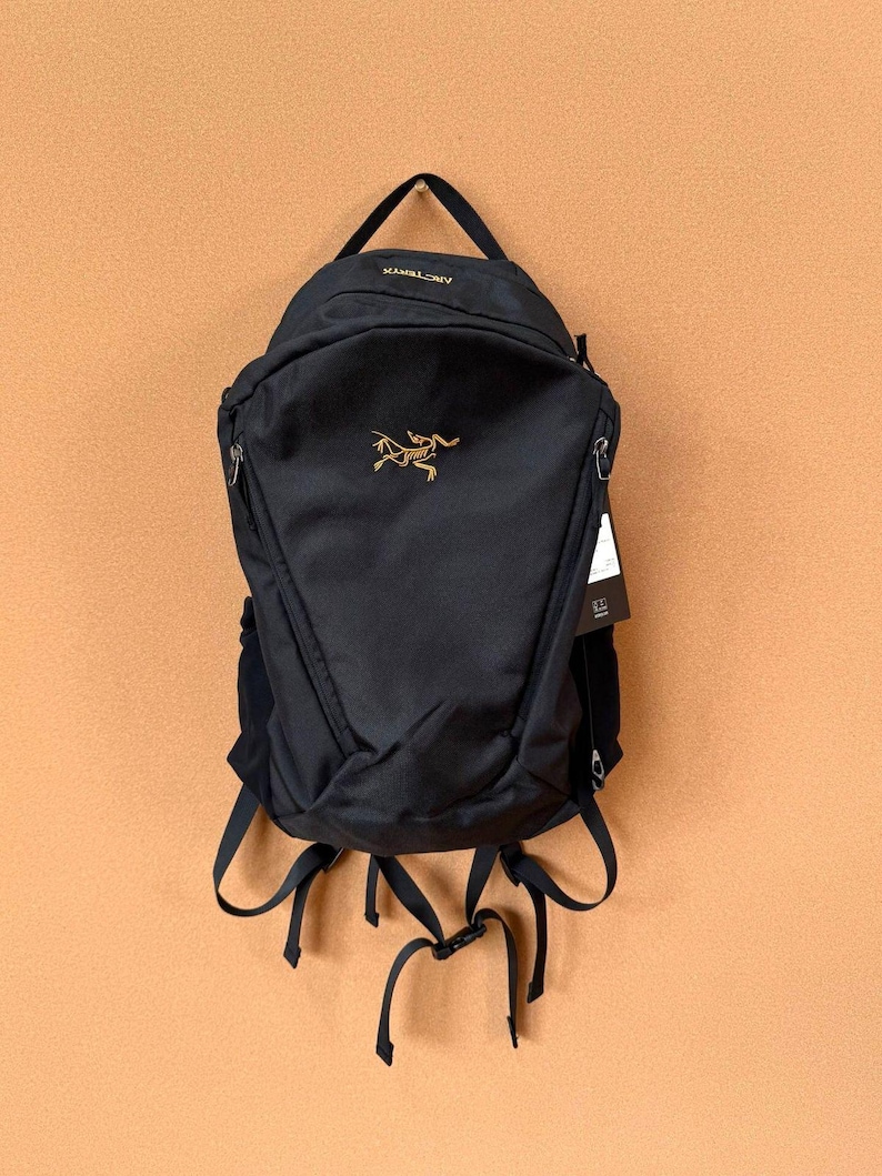 May include: Black backpack with a top handle and adjustable straps. The backpack features a gold logo and the brand name "Arc'teryx" at the top. It has multiple zippered compartments and a sleek, minimalist design.