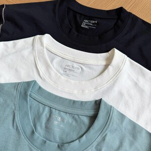 May include: Three crew neck t-shirts in black, white, and teal. Each shirt has a tag with the brand name "ARCTERYX" visible on the collar. The shirts are neatly folded, showcasing their simple design and solid colours. The shirts are made of a soft material.