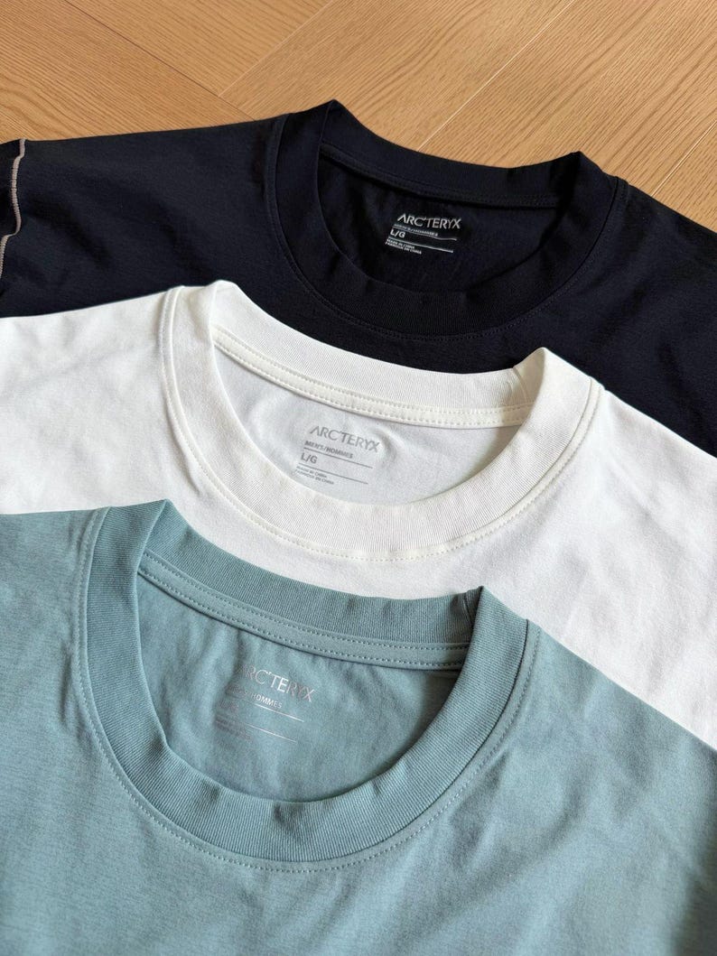May include: Three crew neck t-shirts in black, white, and teal. Each shirt has a tag with the brand name "ARCTERYX" visible on the collar. The shirts are neatly folded, showcasing their simple design and solid colours. The shirts are made of a soft material.