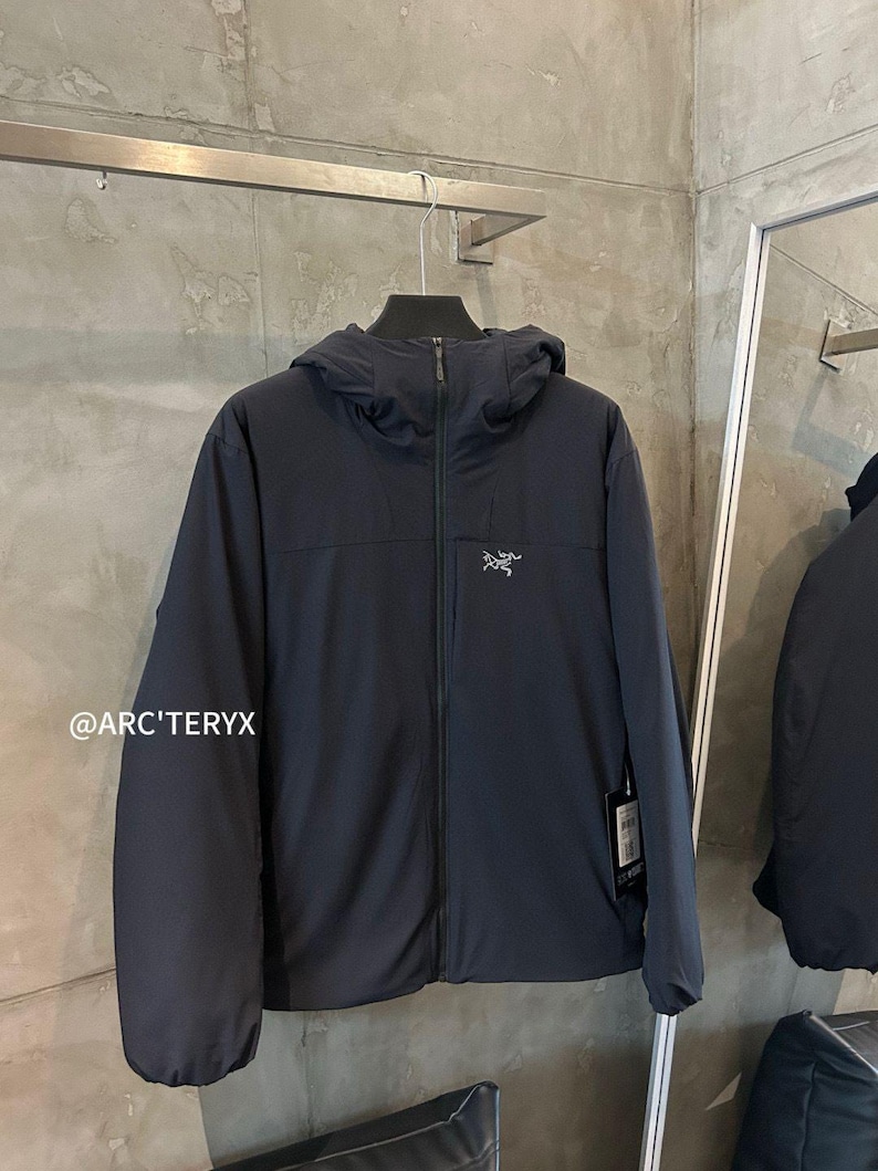 Op de afbeelding: Een marineblauwe jas met capuchon, een ritssluiting over de hele lengte en het Arc'teryx-logo op de borst. De jas hangt aan een metalen rek. De tekst "@ARC'TERYX" is linksonder zichtbaar.