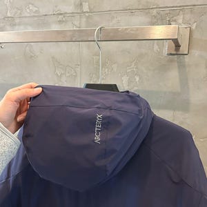 Op de afbeelding: Een marineblauwe jas met capuchon hangt aan een metalen stang. De jas heeft de merknaam "ARC'TERYX" in witte letters op de capuchon. De jas is gemaakt van een glad, mogelijk waterafstotend materiaal.