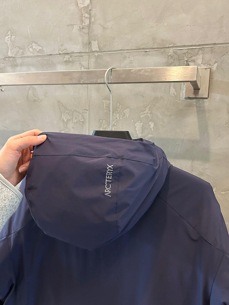 Op de afbeelding: Een marineblauwe jas met capuchon hangt aan een metalen stang. De jas heeft de merknaam "ARC'TERYX" in witte letters op de capuchon. De jas is gemaakt van een glad, mogelijk waterafstotend materiaal.