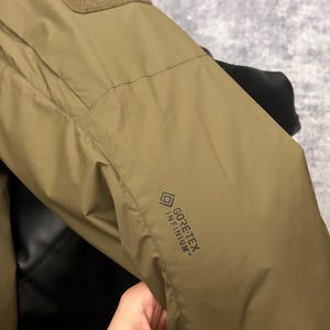 May include: Close-up of a tan jacket sleeve with a rectangular patch and the text "GORE-TEX INFINIUM". The jacket appears to be made of a durable, water-resistant material. The sleeve is slightly wrinkled, suggesting it is flexible.