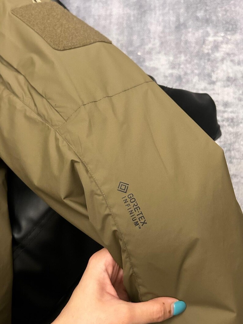 May include: Close-up of a tan jacket sleeve with a rectangular patch and the text "GORE-TEX INFINIUM". The jacket appears to be made of a durable, water-resistant material. The sleeve is slightly wrinkled, suggesting it is flexible.