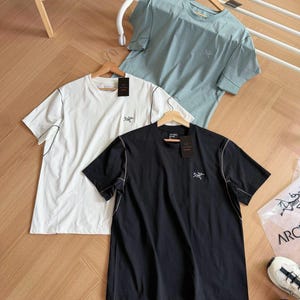 May include: Three short-sleeved t-shirts in white, black, and pale blue, each with a small logo on the chest. The shirts have contrasting stitching and are displayed on wooden hangers. The background includes a wooden floor and a white rack.