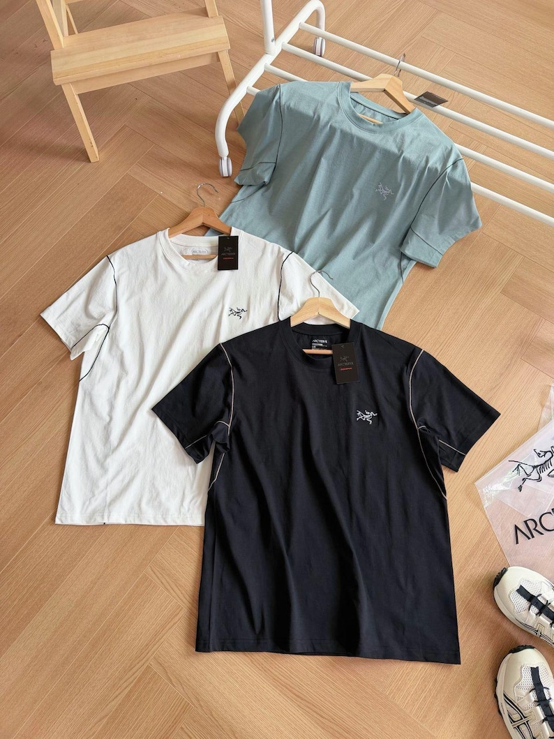 May include: Three short-sleeved t-shirts in white, black, and pale blue, each with a small logo on the chest. The shirts have contrasting stitching and are displayed on wooden hangers. The background includes a wooden floor and a white rack.