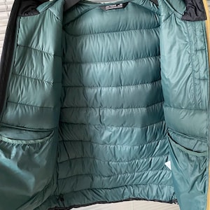 Op de afbeelding: Een zwarte en gouden pufferjack met een teal interieur. De jas heeft een capuchon en twee zakken. De binnenkant is gewatteerd met horizontale stiksels. De merknaam "Arc'teryx" is zichtbaar aan de binnenkant van de jas.