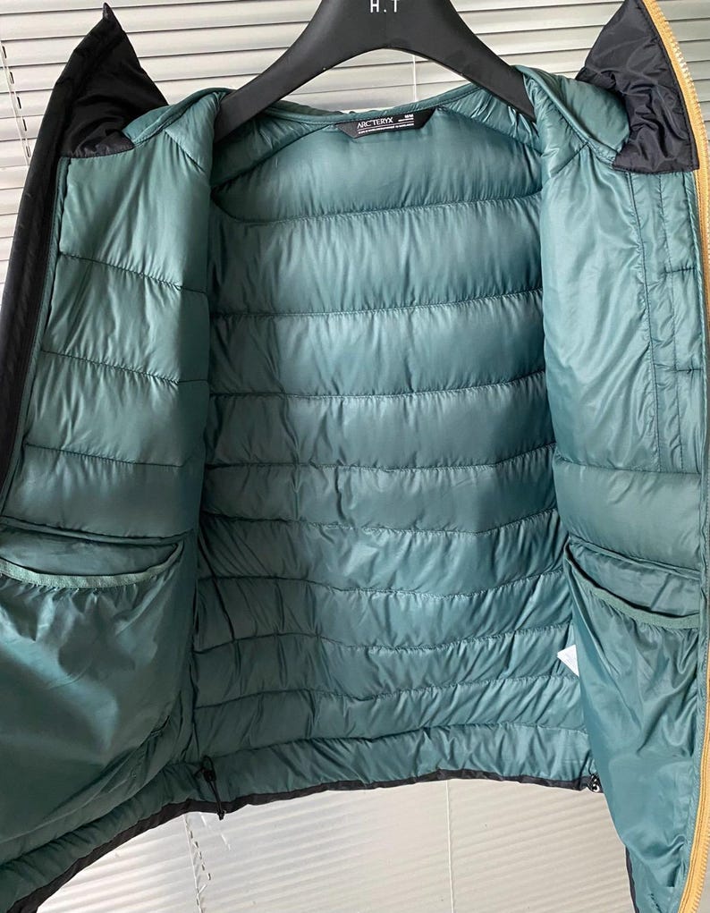 Op de afbeelding: Een zwarte en gouden pufferjack met een teal interieur. De jas heeft een capuchon en twee zakken. De binnenkant is gewatteerd met horizontale stiksels. De merknaam "Arc'teryx" is zichtbaar aan de binnenkant van de jas.