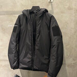 May include: Black hooded jacket with a full-length zipper, two patch pockets, and a small orange pull cord. The jacket has a matte finish and a small black patch on the left sleeve. The brand name "@ARC'TERYX" is visible on the left side.