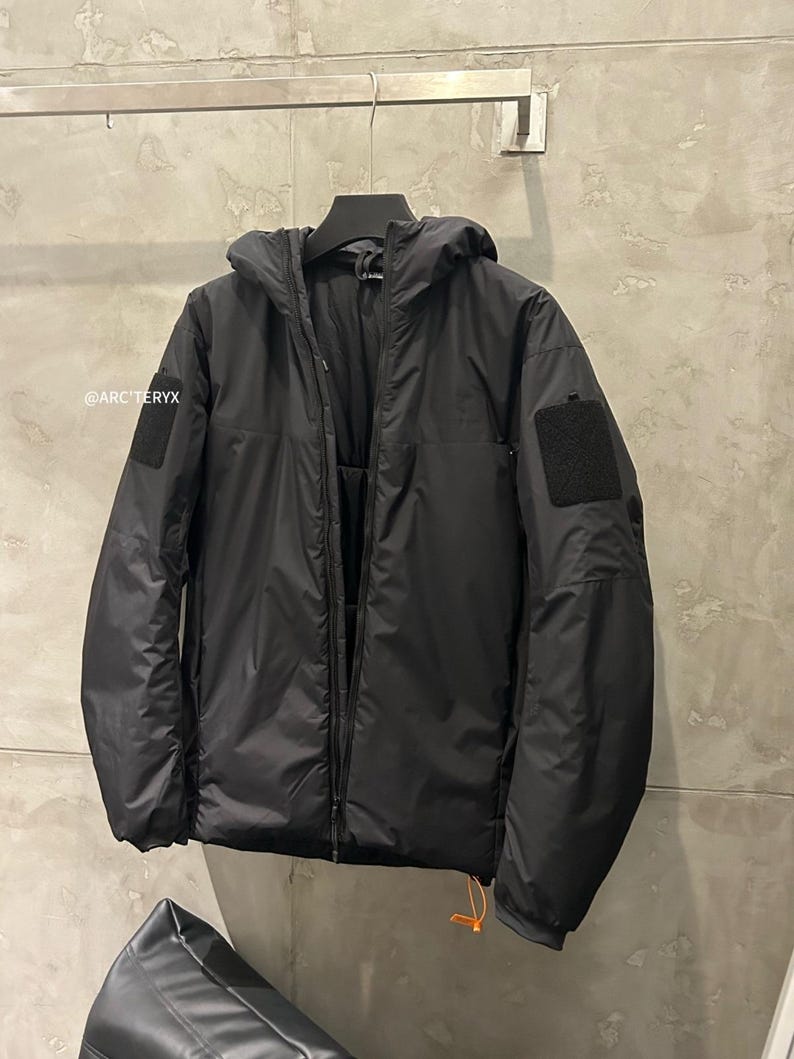 May include: Black hooded jacket with a full-length zipper, two patch pockets, and a small orange pull cord. The jacket has a matte finish and a small black patch on the left sleeve. The brand name "@ARC'TERYX" is visible on the left side.