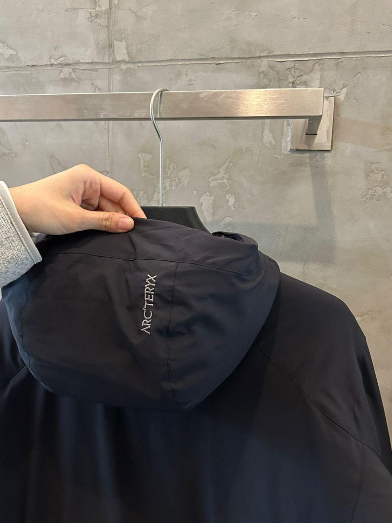 Op de afbeelding: Een marineblauwe jas met capuchon met de merknaam "ARC'TERYX" in witte letters op de capuchon. De jas hangt aan een metalen hanger.