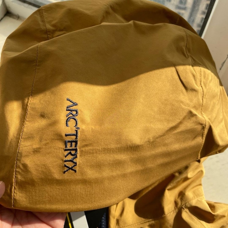 May include: Mustard-coloured hooded jacket with the brand name "ARC'TERYX" embroidered in black on the left side. The jacket appears to be made of a lightweight, water-resistant material. The hood is up, and the jacket is partially visible.