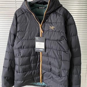 Op de afbeelding: Zwarte gewatteerde jas met capuchon, gouden rits en het Arc'teryx-logo op de borst. De jas heeft twee zakken met ritssluiting en een label. De binnenvoering is groen.