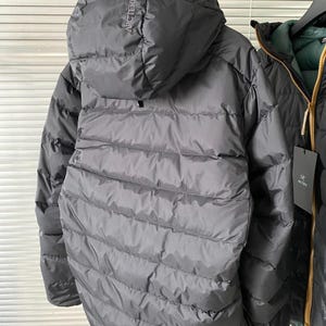 Op de afbeelding: Een grijze donsjas met capuchon met de merknaam "Arc'teryx" op de capuchon. De jas heeft een gewatteerd ontwerp en een ritssluiting over de hele lengte. De jas hangt aan een kledingrek met een andere jas zichtbaar op de achtergrond.