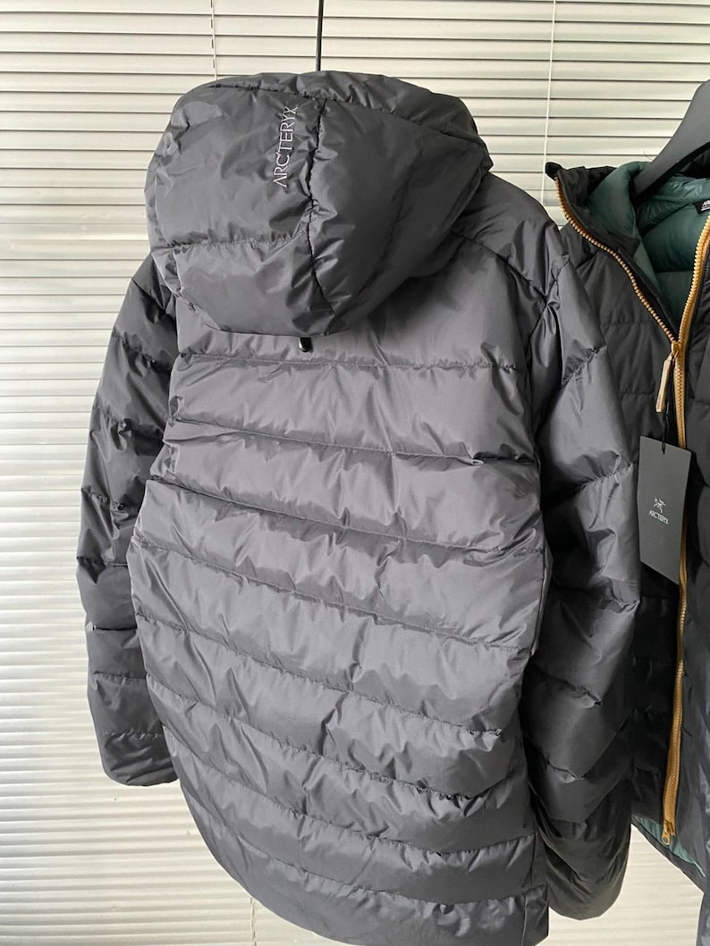 Op de afbeelding: Een grijze donsjas met capuchon met de merknaam "Arc'teryx" op de capuchon. De jas heeft een gewatteerd ontwerp en een ritssluiting over de hele lengte. De jas hangt aan een kledingrek met een andere jas zichtbaar op de achtergrond.