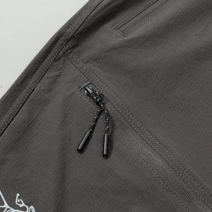 Pode incluir: Close-up de cal&ccedil;as de exterior cinza escuro com uma superf&iacute;cie texturizada. Apresenta um bolso com z&iacute;per com um cord&atilde;o preto e branco e um logotipo branco. As cal&ccedil;as t&ecirc;m um design sob medida.