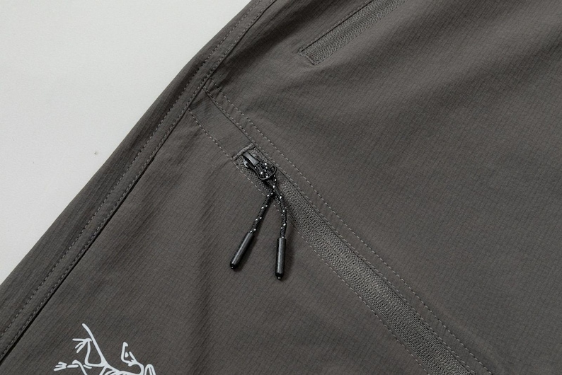 Pode incluir: Close-up de cal&ccedil;as de exterior cinza escuro com uma superf&iacute;cie texturizada. Apresenta um bolso com z&iacute;per com um cord&atilde;o preto e branco e um logotipo branco. As cal&ccedil;as t&ecirc;m um design sob medida.