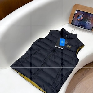 May include: A black puffer vest with a yellow lining, displayed on a white surface. The vest has a zip closure and a small logo on the chest. A tag is attached. The vest is likely designed for outdoor activities.