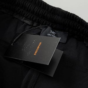 Pode incluir: Close-up de tecido preto com c&oacute;s franzido. Duas etiquetas pretas presas, uma com o logotipo Arc'teryx e a palavra "ENDORPHIN" impressa. O tecido parece ser um material leve.
