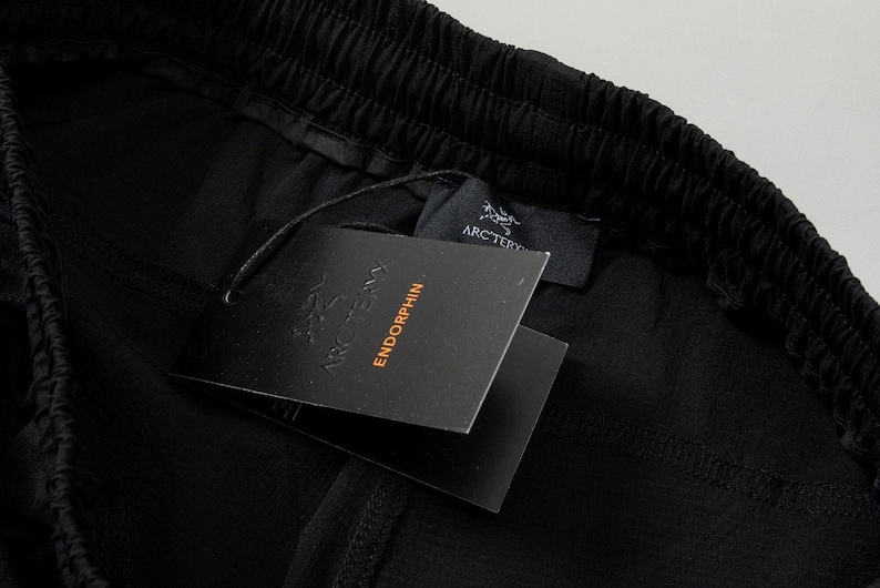 Pode incluir: Close-up de tecido preto com c&oacute;s franzido. Duas etiquetas pretas presas, uma com o logotipo Arc'teryx e a palavra "ENDORPHIN" impressa. O tecido parece ser um material leve.