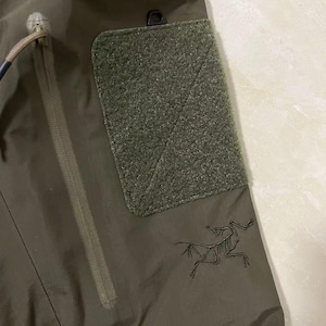 Op de afbeelding: Detail van een olijfgroene tactische broek met een getextureerde patch en een zak met ritssluiting. Voorzien van een zwart geborduurd logo van een skeletvogel. De broek is gemaakt van duurzaam, waterbestendig materiaal.