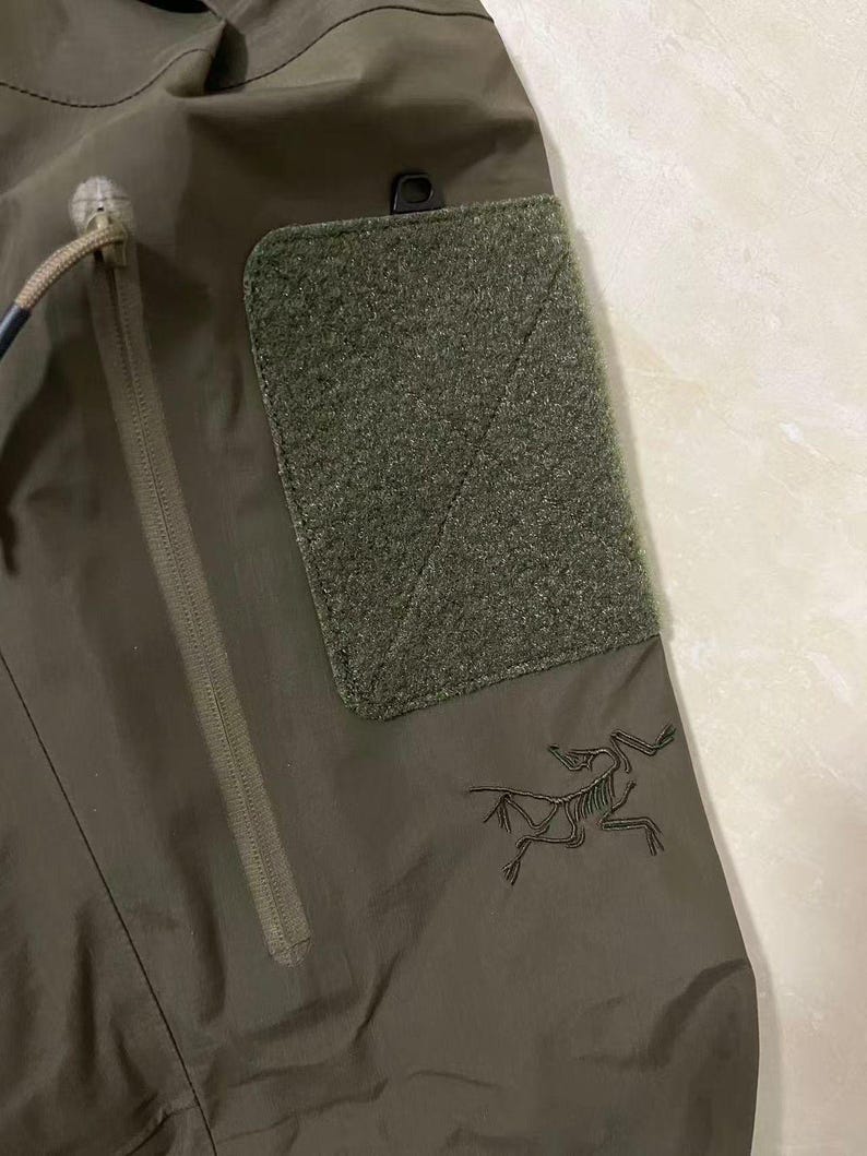 Op de afbeelding: Detail van een olijfgroene tactische broek met een getextureerde patch en een zak met ritssluiting. Voorzien van een zwart geborduurd logo van een skeletvogel. De broek is gemaakt van duurzaam, waterbestendig materiaal.