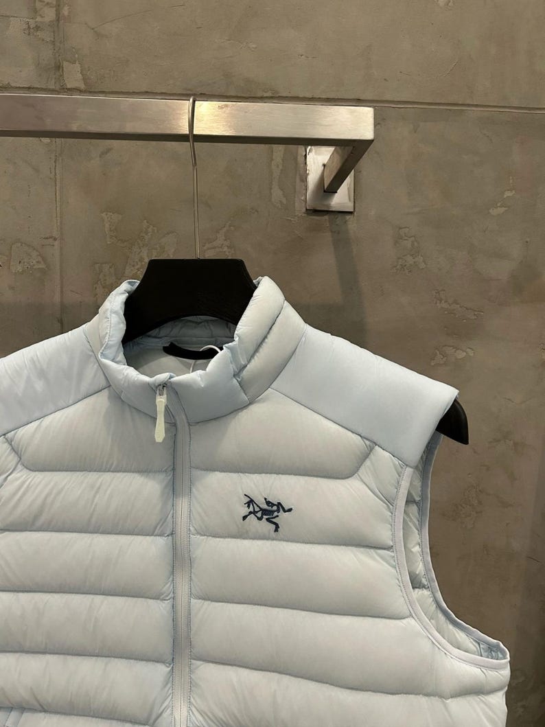 May include: A light blue puffer vest with a high collar and a full-length zipper. The vest features a small black logo on the chest. The vest is hanging on a black hanger against a neutral background.