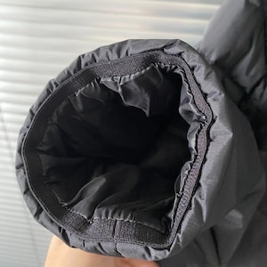 Op de afbeelding: Close-up van een zwarte jas mouwmanchet. De manchet is elastisch en heeft een gewatteerd ontwerp. De binnenkant van de mouw is zwart. De jas lijkt gemaakt van een glad, mogelijk waterafstotend materiaal.