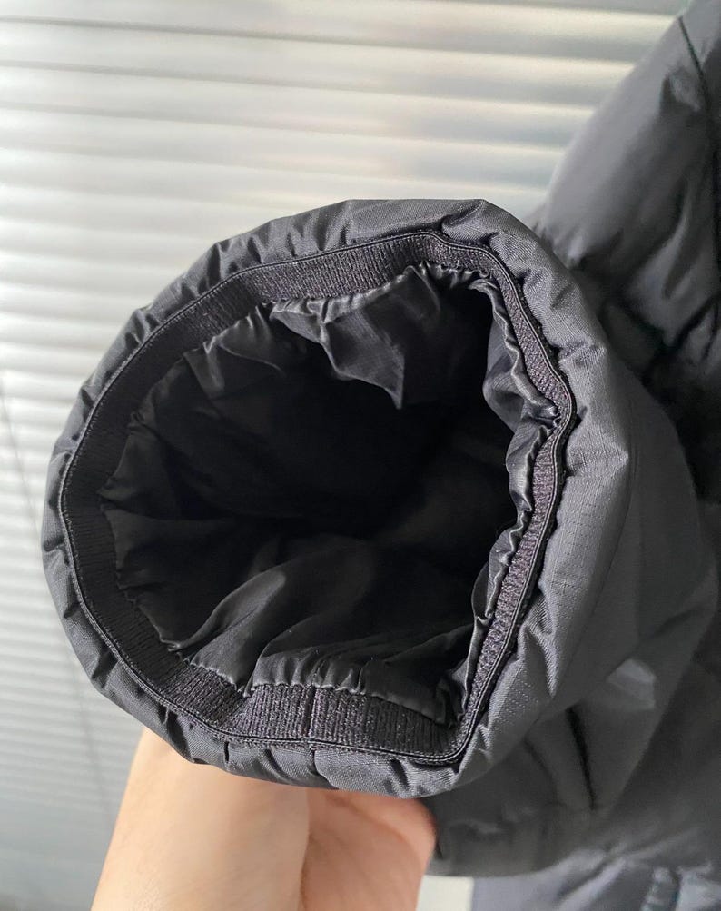 Op de afbeelding: Close-up van een zwarte jas mouwmanchet. De manchet is elastisch en heeft een gewatteerd ontwerp. De binnenkant van de mouw is zwart. De jas lijkt gemaakt van een glad, mogelijk waterafstotend materiaal.