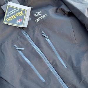 May include: Black Arc'teryx jacket with a GORE-TEX tag. The jacket has two zippered pockets and a full-length zipper. The Arc'teryx logo is on the upper right chest. The GORE-TEX tag is white, yellow, and black.