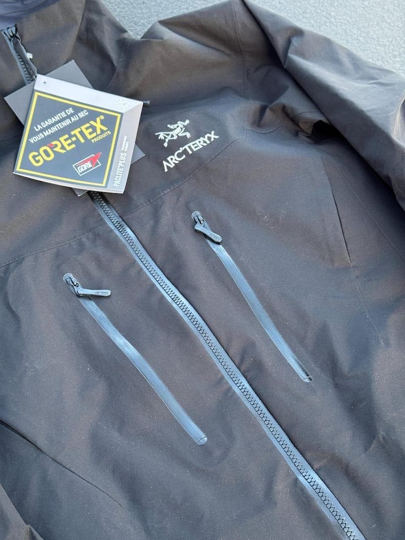 May include: Black Arc'teryx jacket with a GORE-TEX tag. The jacket has two zippered pockets and a full-length zipper. The Arc'teryx logo is on the upper right chest. The GORE-TEX tag is white, yellow, and black.