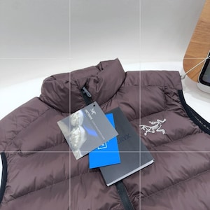 May include: A brown quilted down vest with a high collar and black trim. The vest has a zip closure and a tag with the brand logo. The vest is displayed on a white surface.