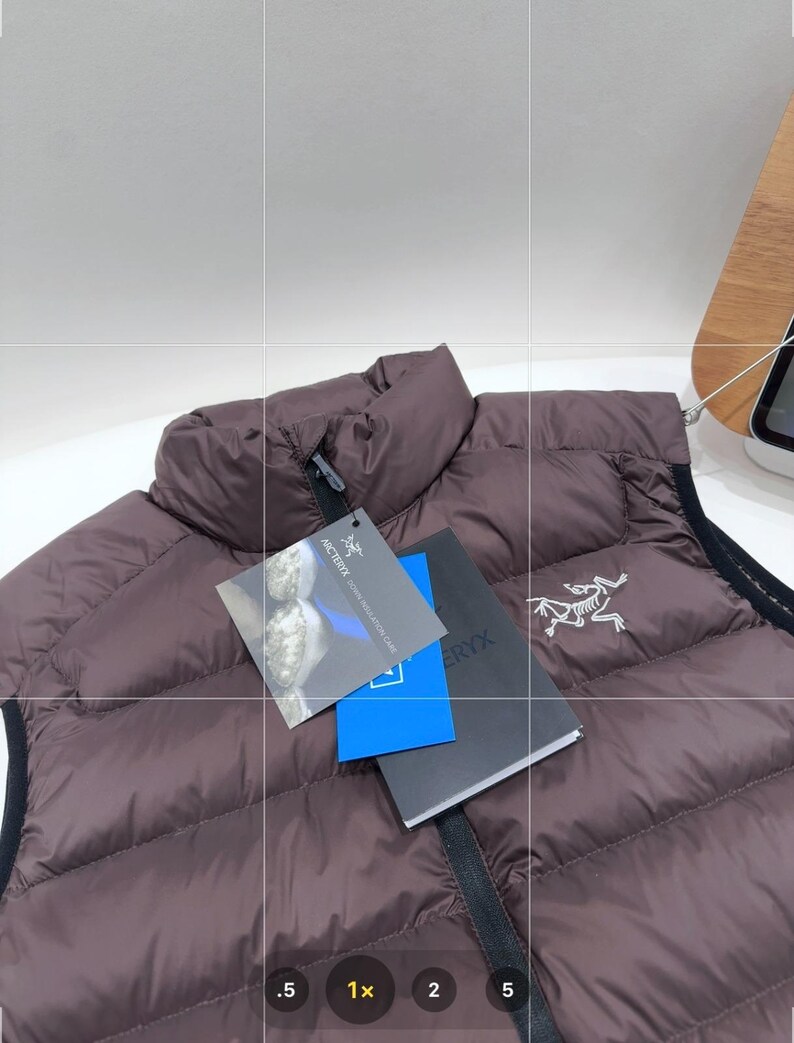 May include: A brown quilted down vest with a high collar and black trim. The vest has a zip closure and a tag with the brand logo. The vest is displayed on a white surface.