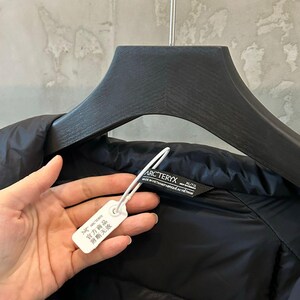 Op de afbeelding: Zwarte gewatteerde jas met een aangehecht label. Het label heeft Chinese karakters en de merknaam. De jas hangt aan een zwarte hanger. De merknaam "Arc'teryx" is zichtbaar op het label.