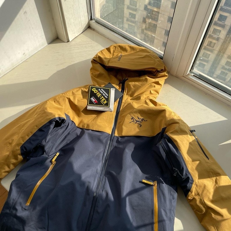 May include: Mustard yellow and navy blue hooded jacket with a black zip and a GORE-TEX tag. The jacket features a small logo on the chest and two zipped pockets. Designed for outdoor activities, it has a practical design.