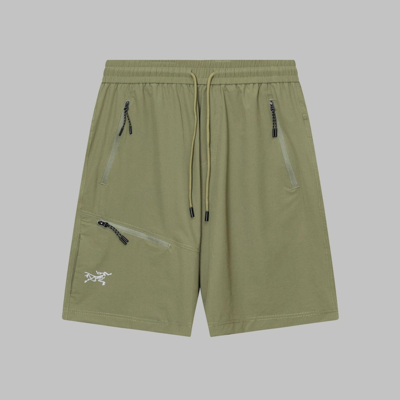 Pode incluir: Cal&ccedil;&otilde;es verde-oliva com cintura com cord&atilde;o e v&aacute;rios bolsos com z&iacute;per. Os cal&ccedil;&otilde;es apresentam um pequeno log&oacute;tipo branco na perna esquerda. O tecido parece leve e adequado para atividades ao ar livre.