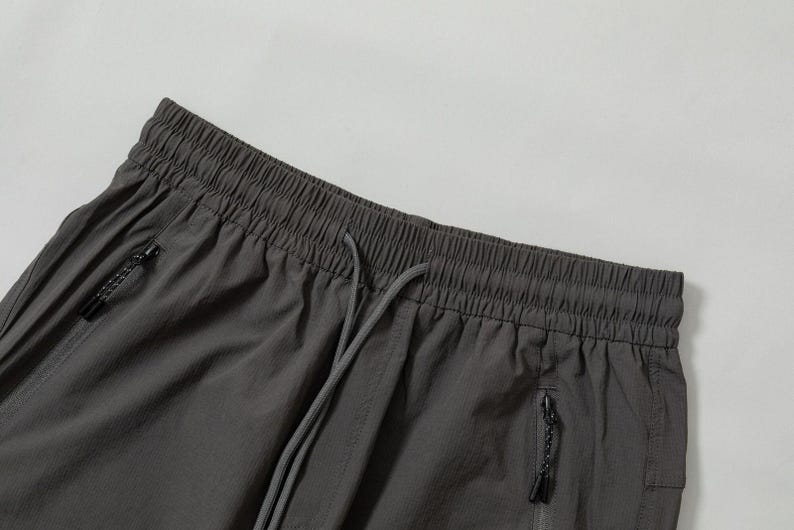Pode incluir: Close-up de cal&ccedil;&otilde;es desportivos cinza carv&atilde;o. Os cal&ccedil;&otilde;es t&ecirc;m uma cintura el&aacute;stica com cord&atilde;o, dois bolsos com z&iacute;per e um pequeno bolso lateral. O tecido parece leve.