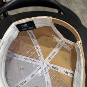 Puede incluir: Gorra de b&eacute;isbol beige con interior blanco y el nombre de la marca "ARC'TERYX" impreso en el interior. La gorra tiene una visera curva y una etiqueta negra con el logotipo de la marca.