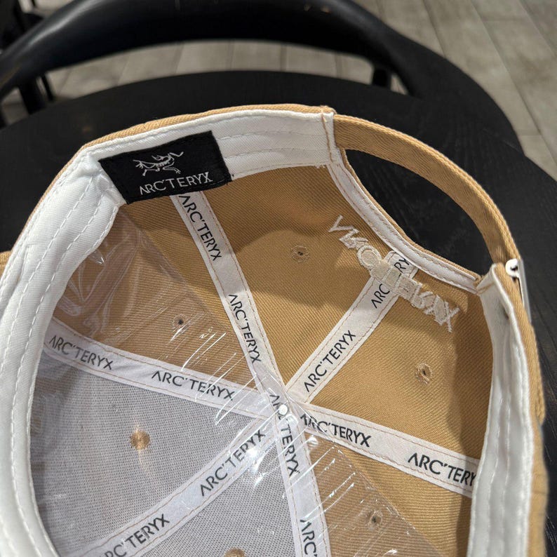 Puede incluir: Gorra de b&eacute;isbol beige con interior blanco y el nombre de la marca "ARC'TERYX" impreso en el interior. La gorra tiene una visera curva y una etiqueta negra con el logotipo de la marca.