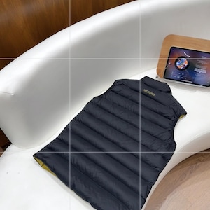 May include: A black quilted vest with a yellow lining, placed on a white surface. The vest features a high collar and the brand name "ARCTERYX" in gold. The vest is made of a lightweight material, possibly designed to be water-resistant.