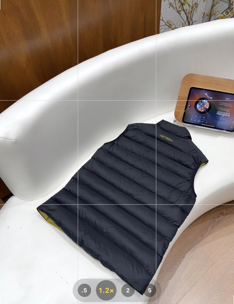 May include: A black quilted vest with a yellow lining, placed on a white surface. The vest features a high collar and the brand name "ARCTERYX" in gold. The vest is made of a lightweight material, possibly designed to be water-resistant.