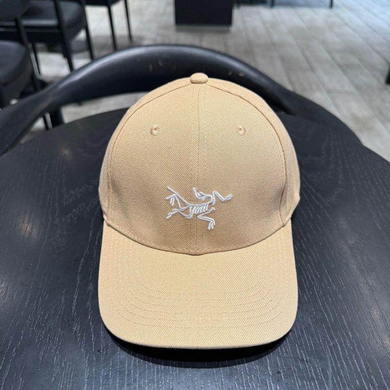 Puede incluir: Gorra de b&eacute;isbol beige con visera curva. La gorra presenta un logotipo blanco bordado en la parte delantera. La gorra est&aacute; hecha de una tela duradera y tiene un dise&ntilde;o cl&aacute;sico de seis paneles. La gorra es de color neutro.