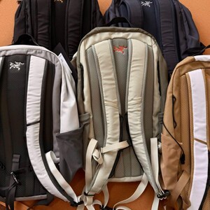May include: A collection of backpacks in various colors, including black, navy, gray, and tan. The backpacks have padded backs and shoulder straps. Each backpack features a logo on the front and back.