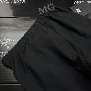 May include: Black outdoor pants with a drawstring waist. The pants are made of a durable, water-resistant material. The Arc'teryx logo is visible on the box in the background.